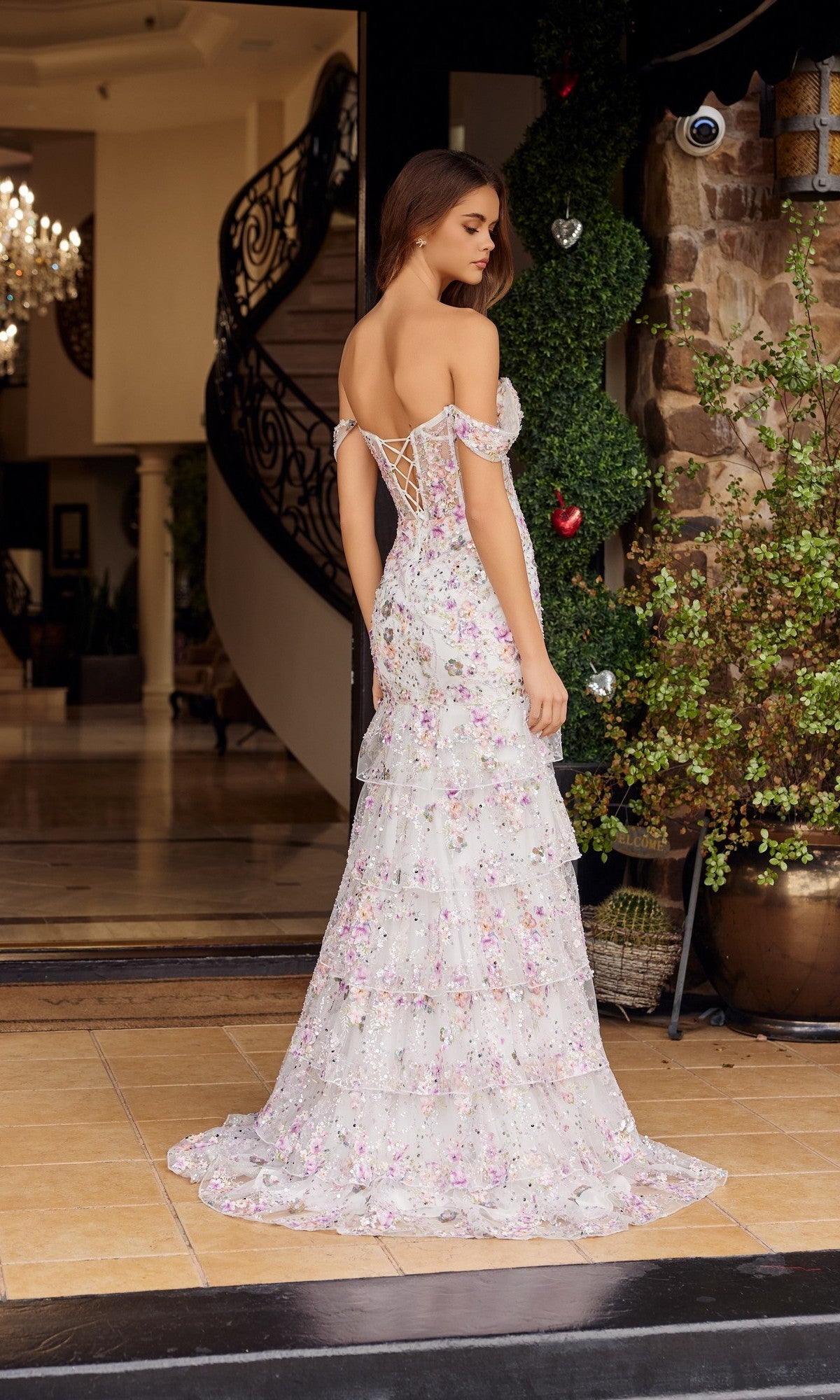A woman wearing the Nox Anabel G1916 Long Prom Dress, a white off-shoulder gown with floral beaded embroidery, stands indoors on tiled flooring near greenery and a spiral staircase, facing away from the camera.